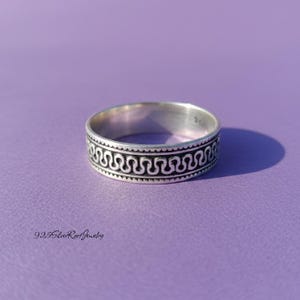 May include: A silver ring with a repeating wave pattern around the band, set against a purple background. The ring's design features a series of connected, curved lines, creating a textured appearance. The ring is a piece of jewelry.