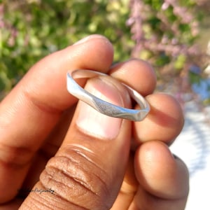 May include: A silver ring with a geometric design. The ring has a faceted surface and a smooth, polished band. The ring is held in a hand against a blurred background of green and purple foliage.