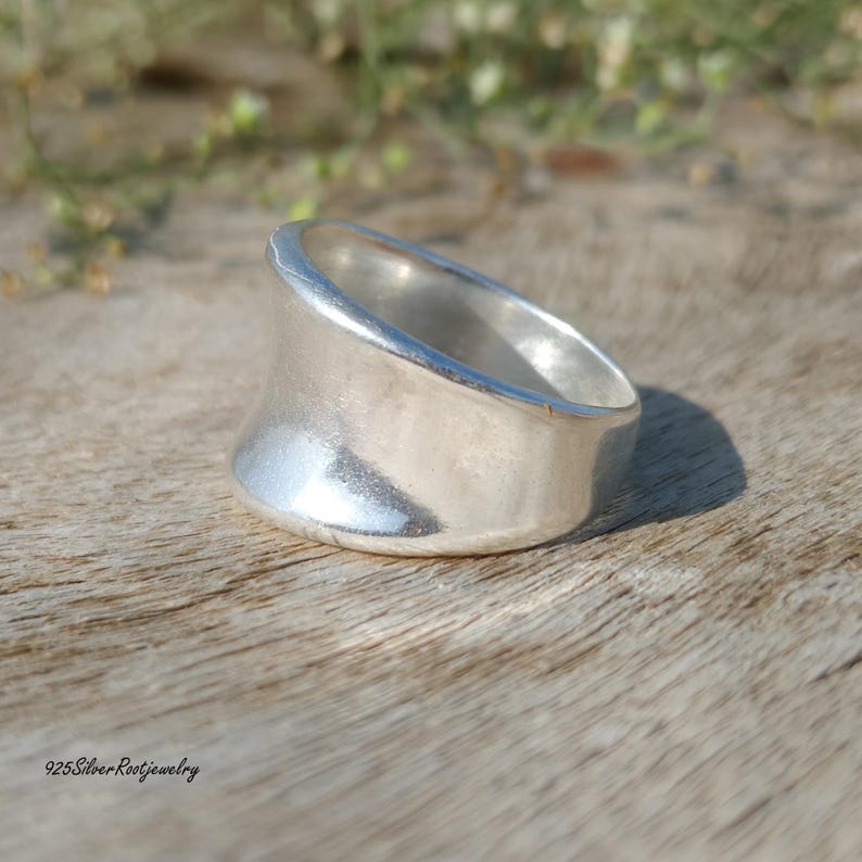 May include: A wide, silver ring with a curved design. The ring has a smooth, polished surface and a thick band. The ring is displayed on a wooden surface. The text "925SilverRootJewelry" is visible at the bottom.