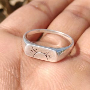 May include: A silver ring with a rectangular face featuring a sunburst design. The ring is made of a shiny, metallic material and is shown in a close-up shot. The sunburst design is etched into the face of the ring.