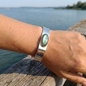 May include: A wide, polished silver cuff bracelet featuring a single, oval-shaped labradorite gemstone set in the centre. The stone displays iridescent green and blue flashes, contrasting with the smooth, reflective metallic surface of the band.