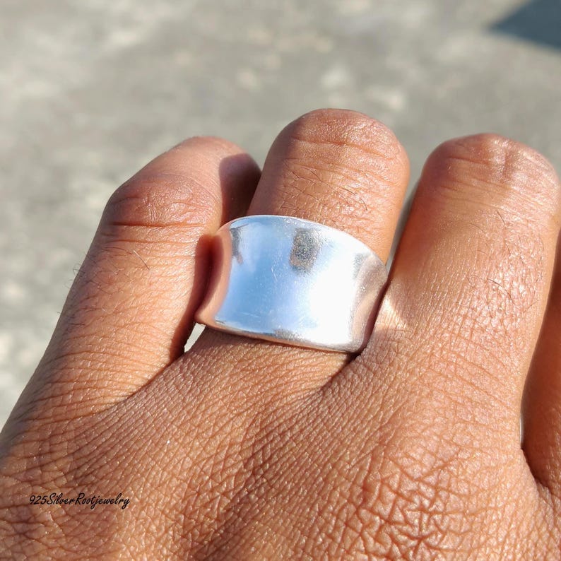 May include: A wide, polished silver ring with a smooth, curved design. The ring is shown on a hand, reflecting light. The jewelry piece has a minimalist style, suitable for everyday wear.