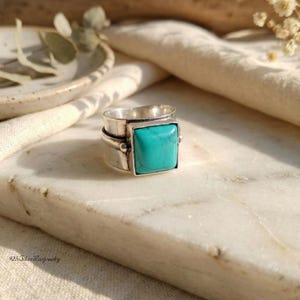 May include: A silver ring with a square, turquoise-colored stone. The ring has a wide band with a textured design and a bezel setting for the stone. The ring is displayed on a marble surface.