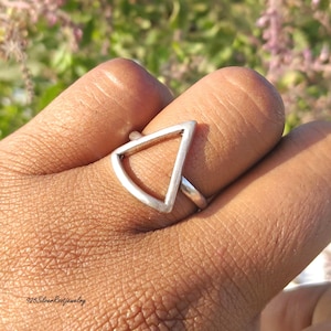 Puede incluir: Un anillo de plata con un diseño geométrico de triángulo. El anillo presenta una banda sólida y una forma de triángulo abierta. El anillo se lleva en un dedo. El fondo está borroso con tonos verdes y morados.