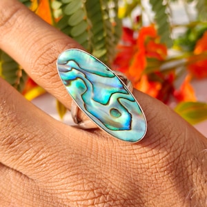 May include: A silver ring featuring an oval abalone shell with iridescent blue, green, and teal hues. The ring's band is a simple, polished silver design. The abalone shell has a unique, swirling pattern.