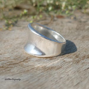 May include: A wide, silver ring with a curved design. The ring has a smooth, polished surface and a thick band. The ring is displayed on a wooden surface. The text "925SilverRootJewelry" is visible at the bottom.