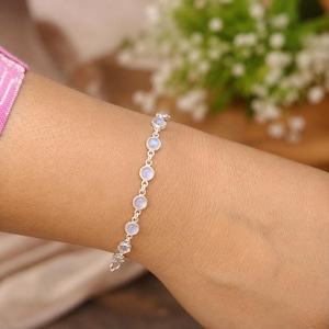 May include: A silver chain bracelet featuring a series of round, bezel-set moonstones. Each stone displays a subtle iridescent blue glow, connected by small circular silver links that form a delicate and continuous line around the wrist.