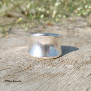 May include: A wide, silver ring with a smooth, polished surface. The ring has a rounded shape and sits on a wooden surface. The text "925SilverRootjewelry" is visible at the bottom left.