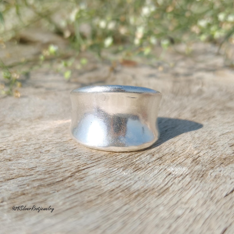 May include: A wide, silver ring with a smooth, polished surface. The ring has a rounded shape and sits on a wooden surface. The text "925SilverRootjewelry" is visible at the bottom left.