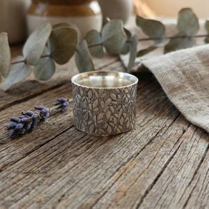 May include: A silver ring with a leaf pattern. The ring is cylindrical and rests on a wooden surface. A sprig of lavender and eucalyptus leaves are in the background. The ring is approximately 2.5 cm in diameter.