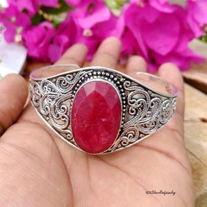 Könnte beinhalten: Ein silbernes Armreif-Armband mit einem ovalen, facettierten, roten Edelstein. Das Armband weist aufwendige, wirbelnde Filigranmuster auf, vor einem Hintergrund aus rosa Blüten.