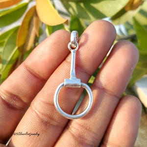 May include: A silver-colored pendant in the shape of a horse bit. The pendant has a circular ring at the bottom, a rectangular center piece, and a vertical bar leading to a loop at the top. The background is blurred with green and yellow foliage.