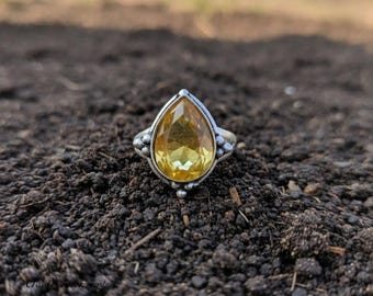 May include: A silver ring with a pear-shaped, yellow gemstone. The ring's band has a decorative design with small, round accents. The gemstone is the focal point, set against a dark, earthy background.