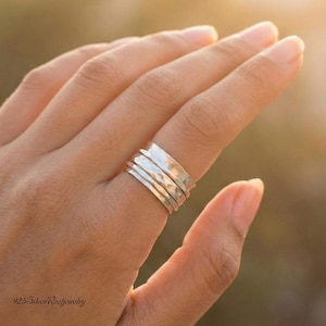 May include: A silver ring with a hammered texture and multiple bands is worn on a finger. The ring has a shiny, metallic appearance. The background is blurred, with warm tones.