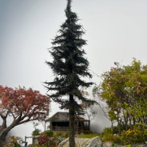 May include: A detailed miniature evergreen tree with dark green foliage. The tree is the focal point, set against a backdrop of a small cabin, other trees, and a cloudy sky. The scene evokes a sense of a miniature landscape or model railway setting.