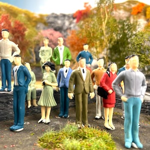 May include: A group of miniature figurines of people in various outfits, including suits and dresses, set in a natural outdoor scene. The background features colourful foliage. The figurines are made of plastic and are posed as if they are in a park.