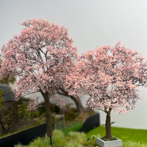 Op de afbeelding: Miniatuurmodelbomen met roze bloesems. De bomen hebben bruine stammen en staan in een graslandschap. De scène omvat een klein gebouw en een grijze lucht op de achtergrond.
