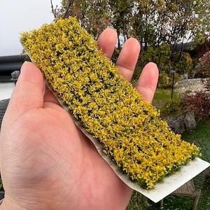 May include: A rectangular model landscape element covered in dense, yellow-green foliage. The miniature plant material is held in a hand, showcasing its texture and color. The base is a white strip, suggesting it's designed for easy placement in a diorama or model scene.
