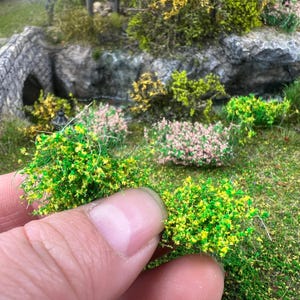 Puede incluir: Primer plano de materiales de modelado de paisajes en miniatura. El primer plano presenta un grupo de arbustos en miniatura de color verde brillante y amarillo. El fondo muestra un puente de piedra en miniatura, rocas y otras plantas en miniatura, creando una escena de diorama detallada.