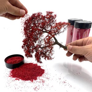 Può includere: Un albero modello in miniatura con un tronco marrone scuro testurizzato e fogliame rosso vibrante. Il set include due barattoli di plastica trasparente con coperchi neri riempiti di materiale floccato rosso e un piccolo contenitore nero con fogliame.
