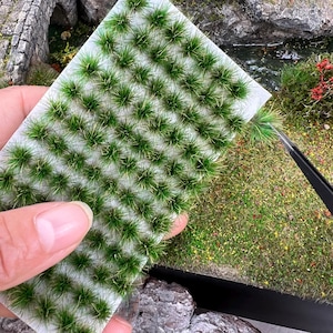 May include: A sheet of miniature green grass tufts, ideal for model landscaping. The grass is a vibrant green with hints of yellow, and is being held by a person's hand. The background shows a miniature landscape with a bridge and other greenery.