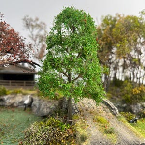 May include: A detailed miniature tree with bright green leaves and a brown trunk. The diorama scene includes other trees with red and yellow foliage, a small pond, and a stone bridge, creating a realistic landscape.