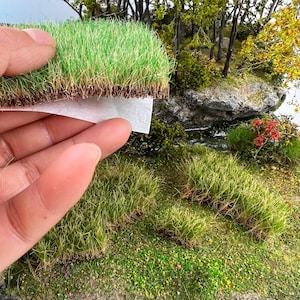 Peut inclure: Gros plan sur une scène de paysage miniature avec une main tenant une section d'herbe artificielle. L'herbe est d'un vert vif avec une base marron, et la scène comprend des arbres, des rochers et d'autres éléments de verdure.