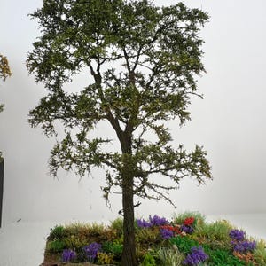 Puede incluir: Un árbol en miniatura con un tronco marrón texturizado y follaje verde. El árbol se asienta sobre una base con flores y hierba en miniatura de colores. El fondo es blanco.