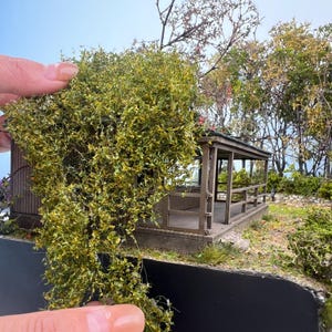 以下が含まれることがあります： 手すりと屋根が付いたミニチュアの木製ポーチ。詳細なモデルの木と葉で囲まれています。このシーンには、小さな暗い道路と木の背景が含まれており、現実的な屋外環境を作り出しています。