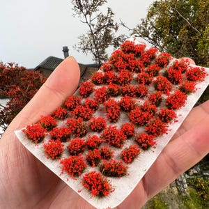 Puede incluir: Una colección de arbustos en miniatura con flores rojas sobre una base blanca. Las pequeñas flores rojas vibrantes están agrupadas, creando una apariencia densa y texturizada. Estas plantas modelo se utilizan probablemente para proyectos de manualidades o modelismo.
