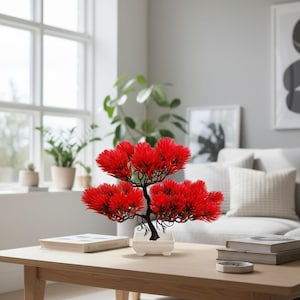 May include: A decorative bonsai tree with vibrant red foliage and a dark trunk sits in a white rectangular pot. The artificial plant is displayed on a light wood coffee table in a living room setting. Books and a small dish are also on the table.