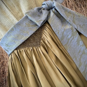 May include: Close-up of a formal dress in olive green with a decorative bow. The bow is a light blue with a subtle pattern. The dress has a pleated skirt and a detailed bodice with small embellishments. The background is a neutral color.