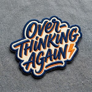 May include: Embroidered patch with the words "Over Thinking Again" in blue and orange lettering. The patch has a white background and a blue border. A small orange lightning bolt is on the right side.