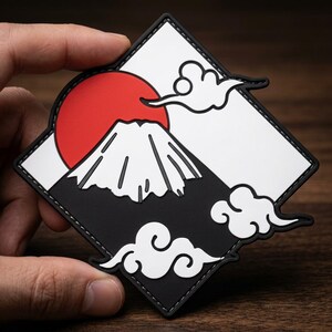 May include: A diamond-shaped PVC patch featuring a graphic design of a white mountain peak against a solid red sun. The background is divided into white and black sections with stylized white clouds. The patch has a black border with stitching.
