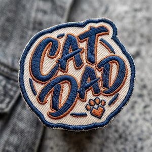 May include: Embroidered patch with the words "CAT DAD" in navy blue and orange thread. The patch has a light beige background and a navy blue border. A small orange paw print is at the bottom.