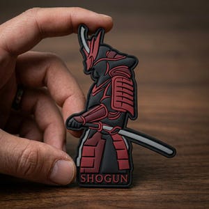 May include: A black and dark red rubber patch featuring a stylized samurai figure in traditional armor with a helmet and a sword. The figure is depicted in profile, and the word SHOGUN is printed in bold red capital letters at the bottom base.