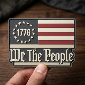 May include: Rectangular patch featuring a stylized American flag design. The upper left corner displays the year "1776" surrounded by stars. The flag's stripes are red and white, and the text "We The People" is displayed below.