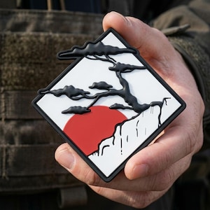 May include: A square-shaped morale patch featuring a raised black relief of a stylized bonsai tree set against a white background. A large, solid red circle representing the sun is positioned behind the tree, with a black border framing the design.