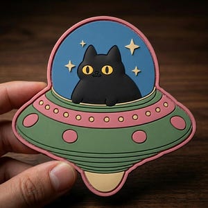 May include: A whimsical patch featuring a black cat inside a UFO. The cat has yellow eyes and is set against a blue background with stars. The UFO is green and pink, with a pink border. A fun, cartoonish design.