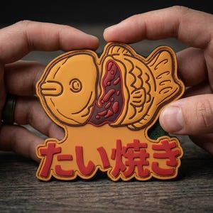 May include: A rubberized patch shaped like a taiyaki fish pastry with a golden-yellow exterior and a dark red bean filling visible in the center. The Japanese text "たい焼き" is embossed in red at the bottom of the fish-shaped design.