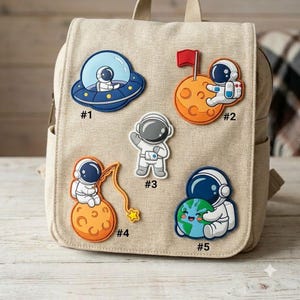 May include: A beige canvas backpack featuring five space-themed patches. Patch #1 shows an astronaut in a UFO. Patch #2 depicts an astronaut on a moon with a red flag. Patch #3 is a standing astronaut. Patch #4 shows an astronaut fishing on a moon. Patch #5 features an astronaut hugging Earth.
