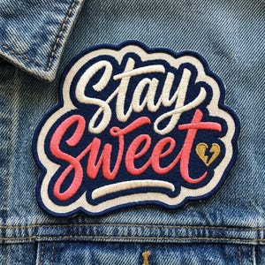 May include: Embroidered patch with the words "Stay Sweet" in white and pink script on a navy blue background. The patch has a white border and a gold heart with a lightning bolt. The patch is on a denim jacket.