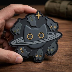 May include: A black patch featuring a gray cat with gold eyes, surrounded by gray ghosts and a planet ring. A gold star is in the upper right corner. The patch is held in a hand.