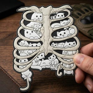 May include: A black and white sticker featuring a rib cage design filled with cartoon cats. The rib cage is beige, and the cats are white with black outlines. The sticker is held by a hand.