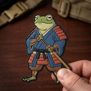 May include: A rubberized patch featuring a green frog dressed in blue and red samurai armor. The frog is depicted standing upright, wearing a dark blue kimono with red shoulder and hip plates, a black belt, and holding a katana sword diagonally.