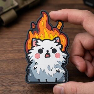 May include: A patch featuring a cartoon cat engulfed in flames. The cat has white fur, pink cheeks, and a surprised expression. The flames are in shades of orange, yellow, and red, outlined in black.