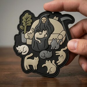 May include: A black patch featuring a Grim Reaper character holding a cat, surrounded by several other cats. The Reaper is seated on a beige couch with a scythe behind it. A small potted plant is also present. The patch has a black border.