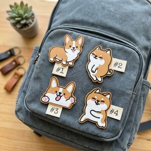 May include: Four rubberized dog-themed zipper pulls attached to a gray canvas backpack. Each charm features a cartoon dog design with a small white tag labeled #1, #2, #3, or #4. The designs include two standing corgis and two Shiba Inu-style dogs.