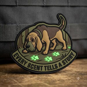 May include: A tactical morale patch featuring a brown and tan bloodhound dog tracking a scent. The design includes glowing green paw prints on the ground and the text EVERY SCENT TELLS A STORY in black lettering against an olive green background.