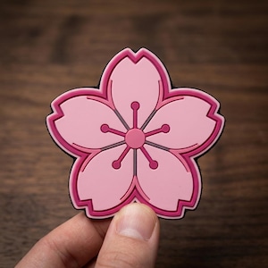 May include: A five-petaled cherry blossom patch in shades of light and dark pink. The flower features a central circular stamen with five radiating lines ending in small circles, all outlined in a bold dark pink border against a black backing.